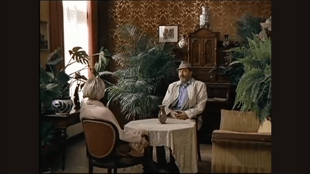 Robert Liebling und Isolde Isenthal sitzen in der Mitte des Bildes an einem Runden Tisch. Sie befinden sich in einer antik aussehenden Kanzlei. Der Raum ist voll mit verschiedenen, ausladenden Zimmerpflanzen.